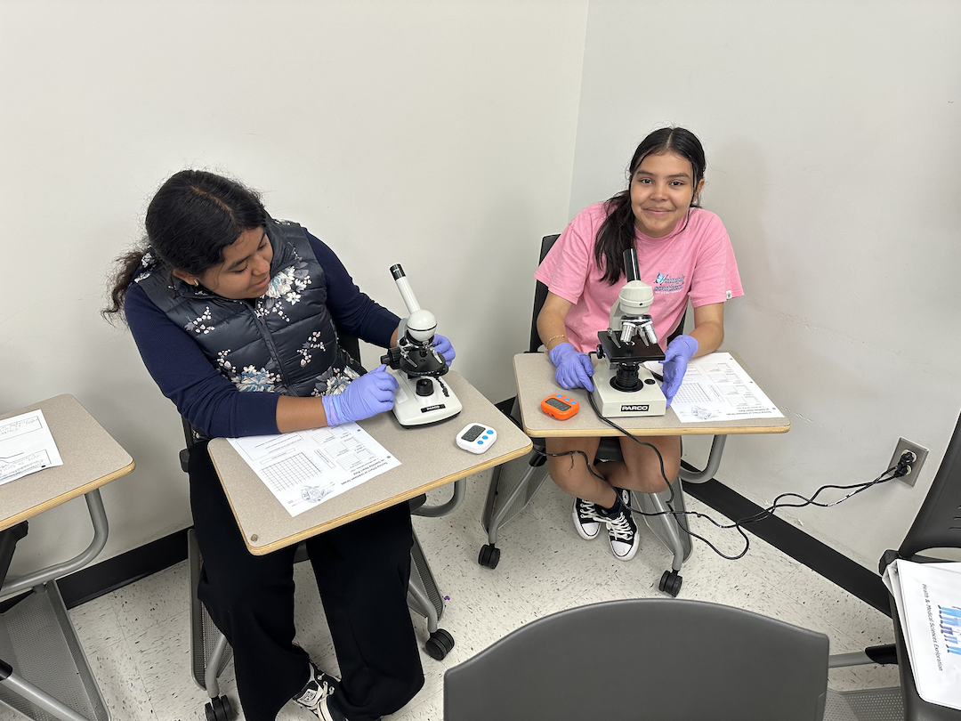 Two middle school aged scholars sit at desks with microscopes.