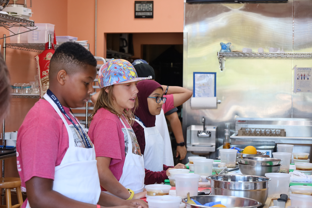 Three middle school aged students standing along a table with white aprons on, preparing food.